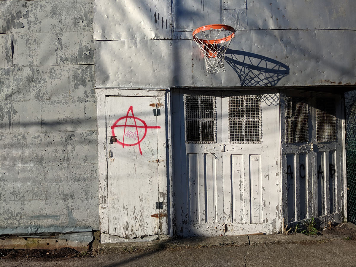 Anarchy hand drawn on old door in alley sided garage with non printed in lighter and smaller text, basketball net with late sun shadows