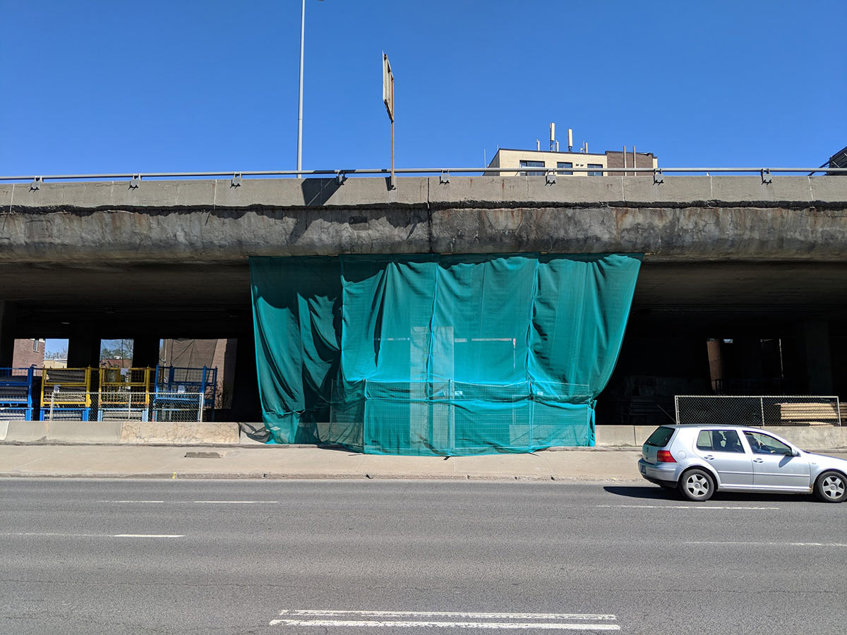 walking along cremazie on a sunny day, veiled mass construction under crumbling cement overpass, car passes by